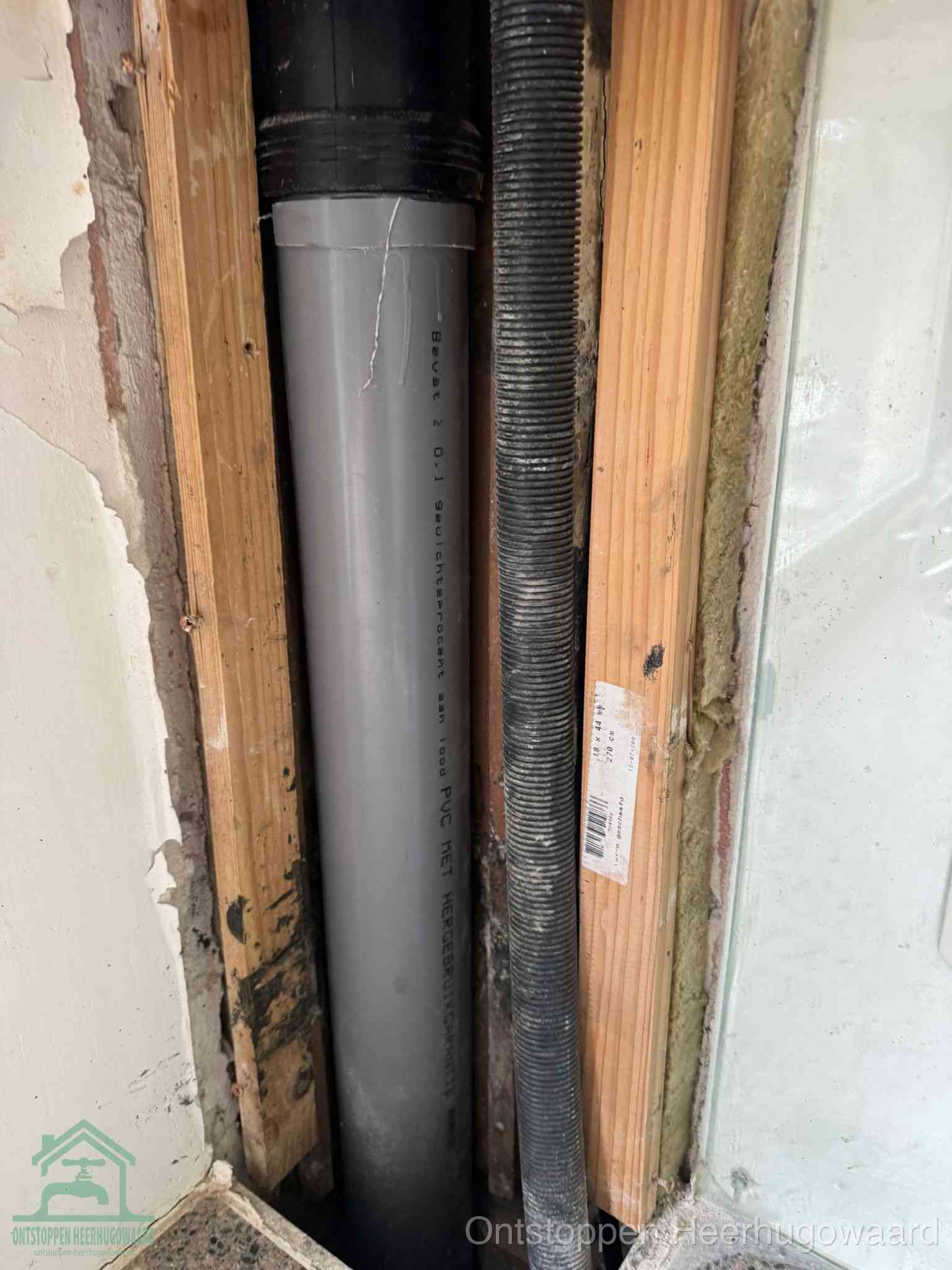 Een grijze PVC-afvoerbuis met een flexibele ribbelslang in een open wandconstructie, een verwaarloosde afvoer die te lang is blijven zitten in Heerhugowaard