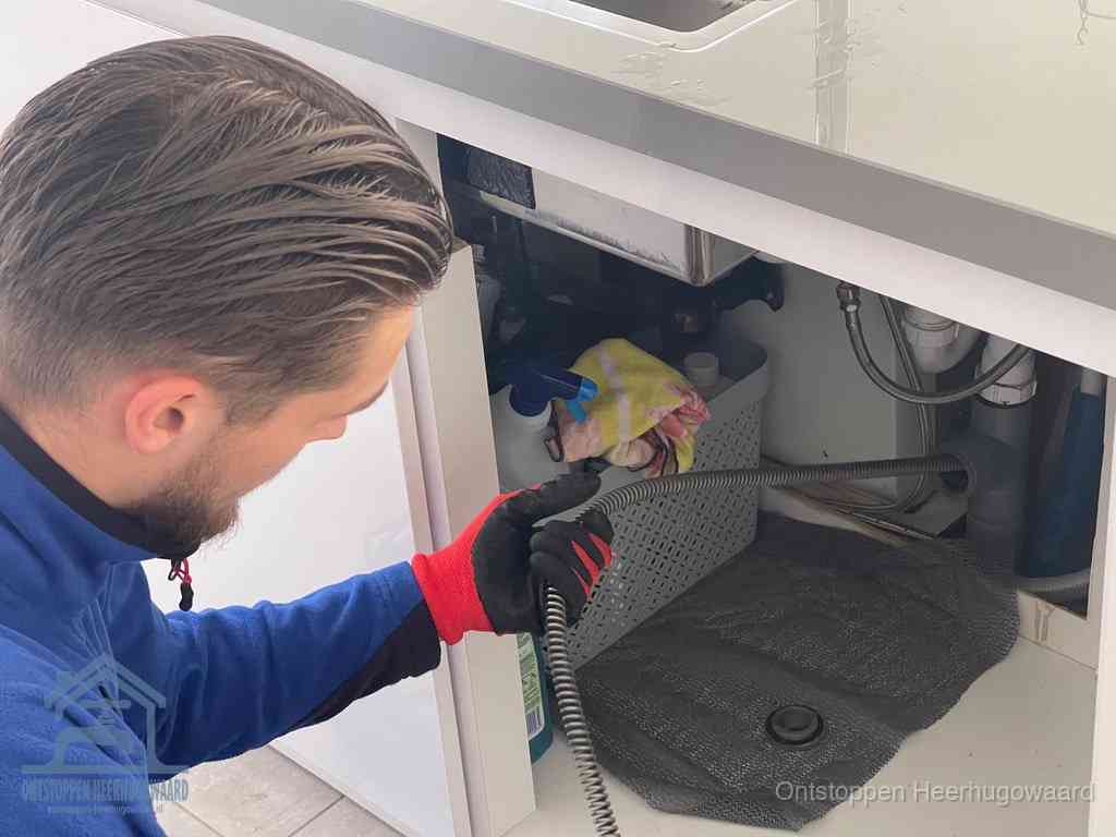 Een monteur voert een flexibele ontstoppingsveer in onder het keukenkastje, langs de sifon, om de gootsteen te ontstoppen in Heerhugowaard