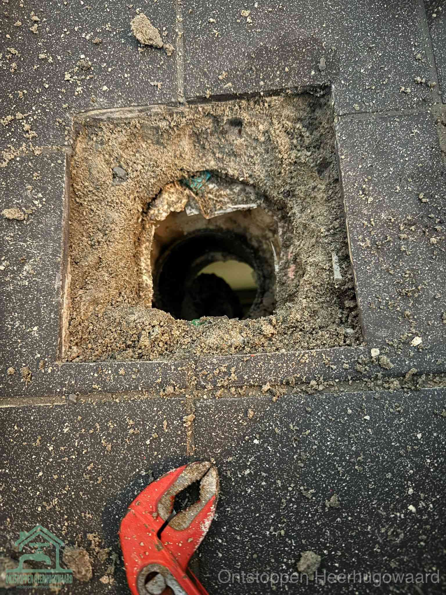 Een geopend rioolputje in een betontegelvloer met losgemaakt rooster en een zichtbare ronde afvoerbuis, het achterhalen waar de afvoerverstopping zit in Heerhugowaard
