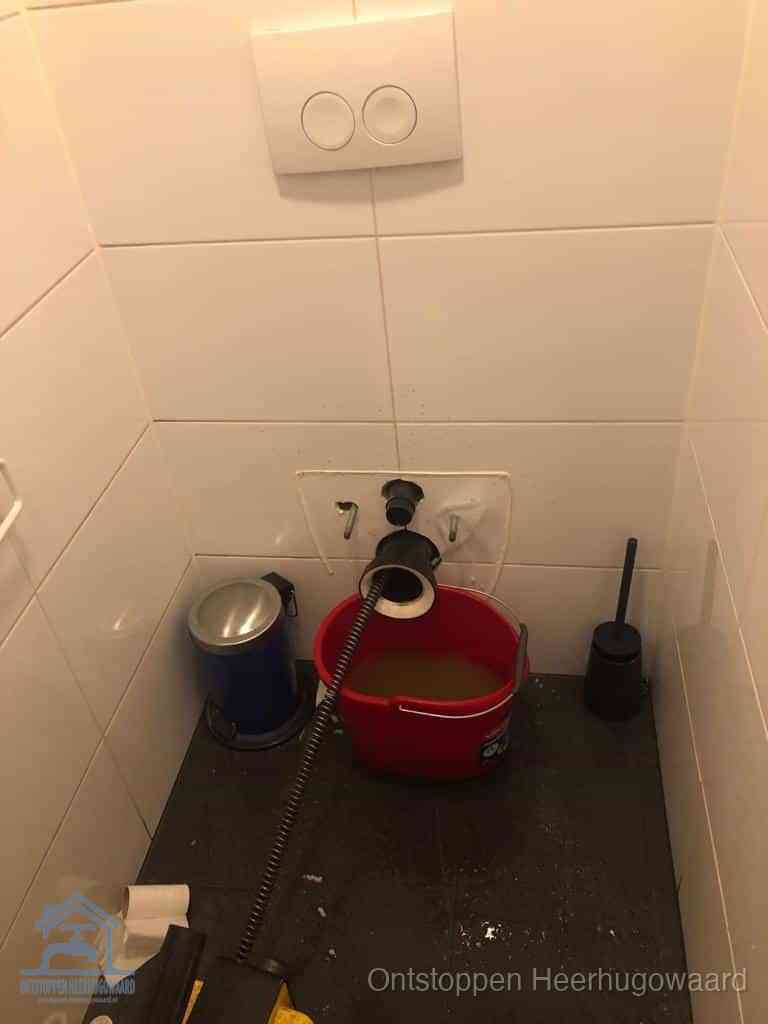 Een ontstopte wand-wc met verwijderde toiletpot waar een ontstoppingsveer in de wanduitgang verdwijnt, het achterhalen waar de verstopping zit in Heerhugowaard