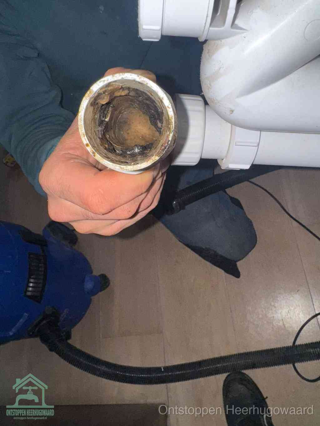 Een hand houdt een losgekoppeld PVC-afvoerbuisstuk vast dat van binnen volledig is dichtgeslibd met vet, vuil en aanslag, de meest voorkomende oorzaak van een verstopte afvoer in Heerhugowaard