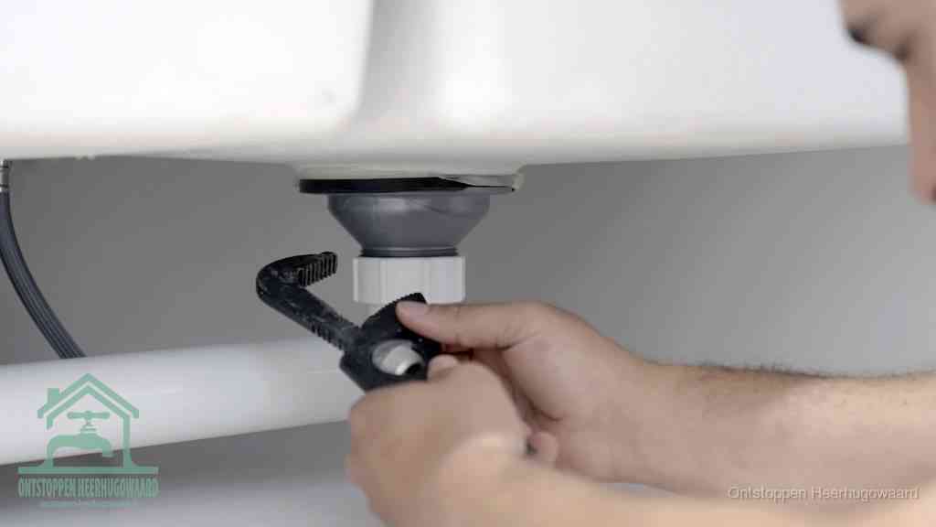 Twee handen draaien met een waterpomptang de wartelmoer van een kunststof sifon onder een wastafel los om de sifon schoon te maken in Heerhugowaard