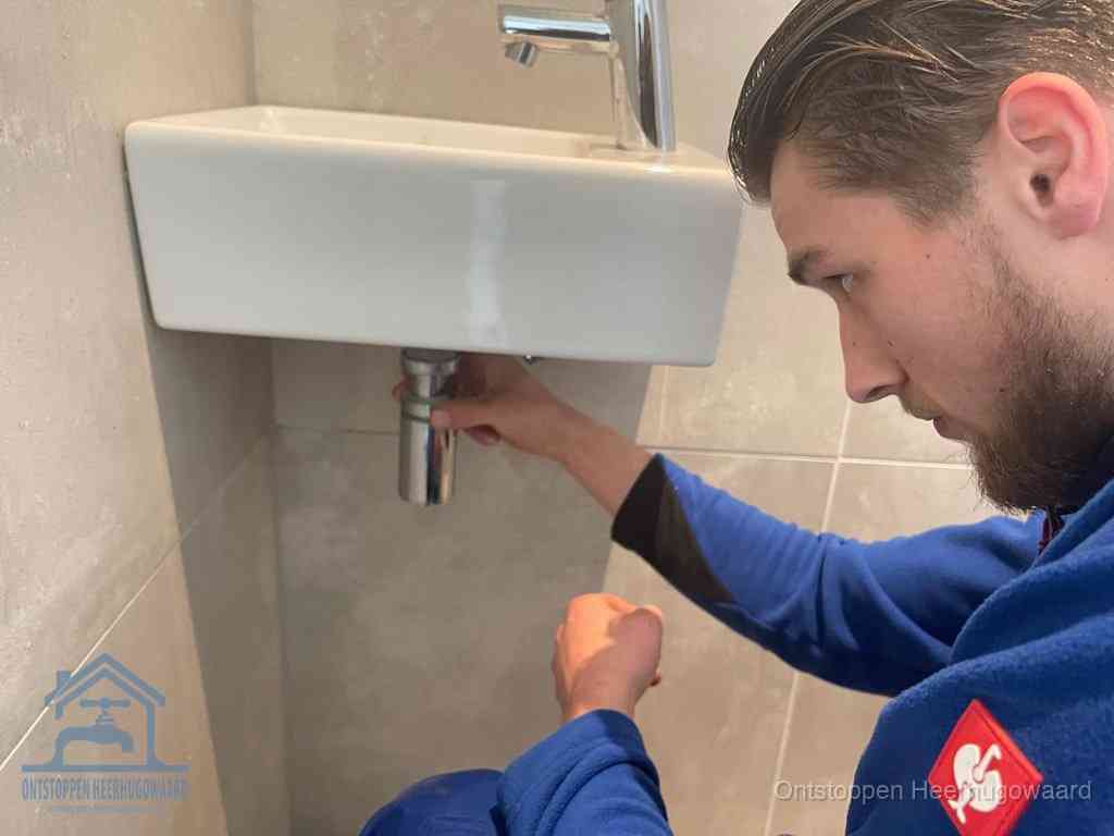 Een monteur draait met de hand de zwanenhalssifon onder een fonteinwasbak in de toiletruimte los om de bron van rioollucht in Heerhugowaard te vinden