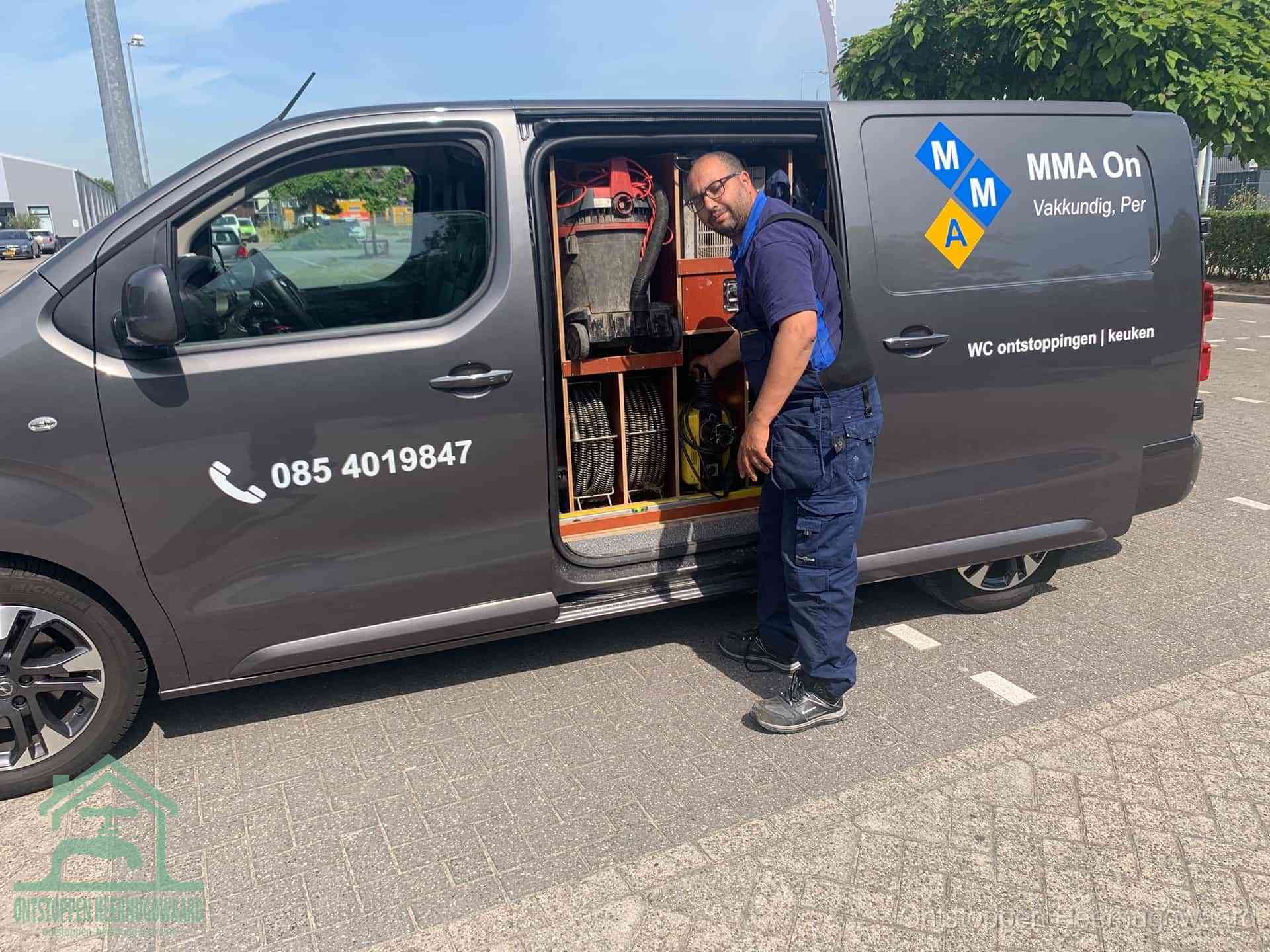 Een monteur bij de open schuifdeur van zijn bedrijfsbus op een parkeerplaats, klaar om de oorzaak van terugkerende afvoerverstoppingen aan te pakken in Heerhugowaard