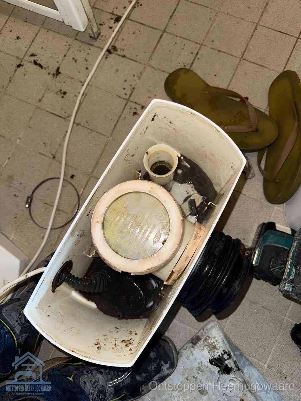 Een gedemonteerde toiletpot op de vloer met ontstoppingsveer en gereedschap ernaast, het verwijderen van een vastzittend voorwerp uit de wc in Heerhugowaard