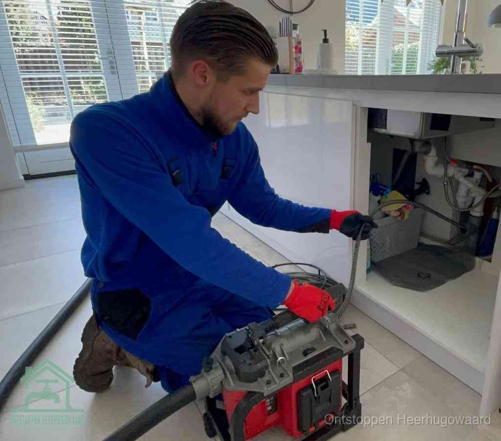 Een monteur met rode handschoenen knielt in een keuken en voert een elektrische ontstoppingsveer in tijdens het professioneel afvoer ontstoppen in Heerhugowaard